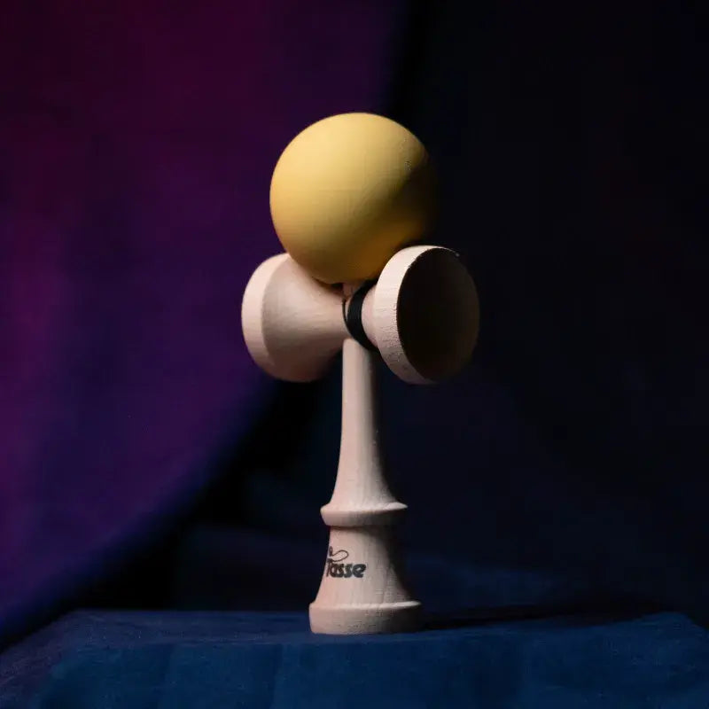 Tasse wooden kendama with a pale yellow ball and natural wood handle, displayed upright on a dark purple and blue background.