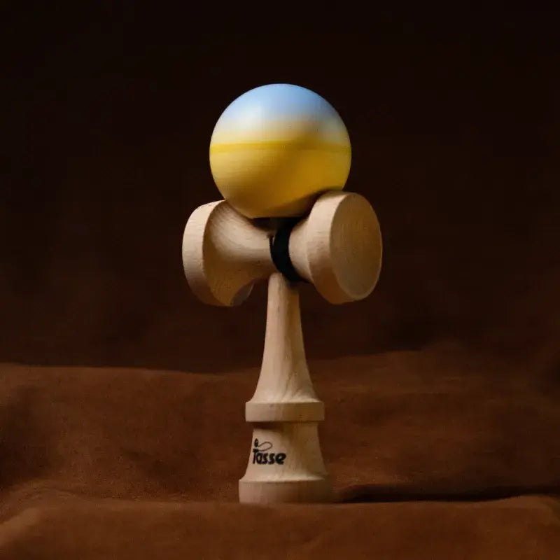 Tasse wooden kendama with a light blue and yellow gradient ball and natural wood handle, displayed upright on a warm brown background.