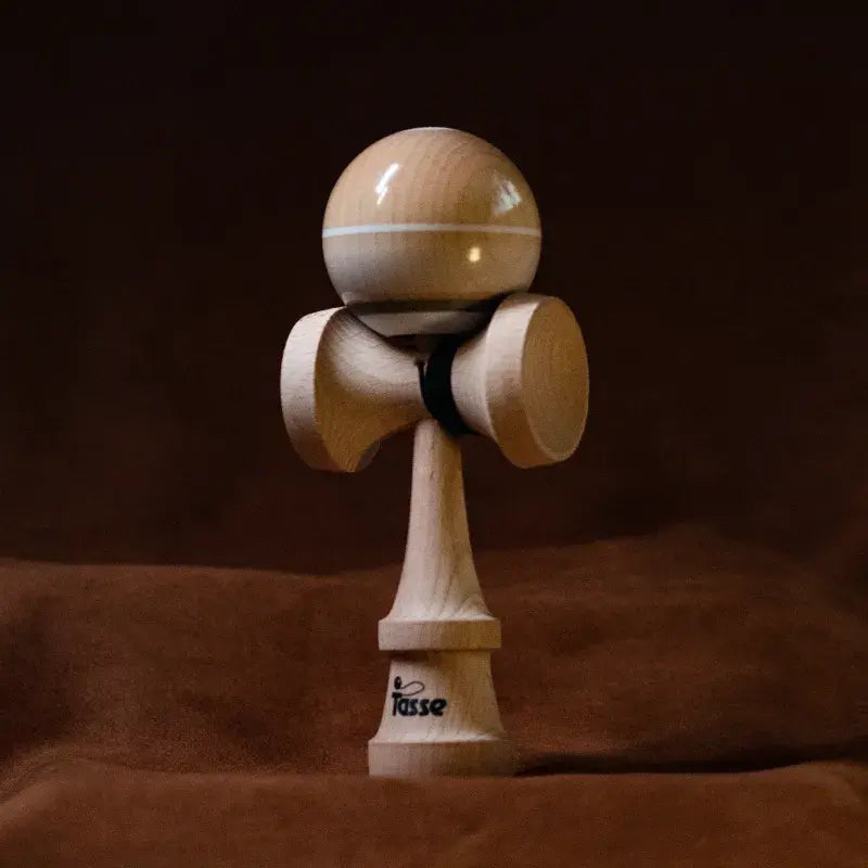 Tasse wooden kendama with a glossy natural wood ball, white stripe detail, and natural wood handle, displayed upright on a warm brown background.