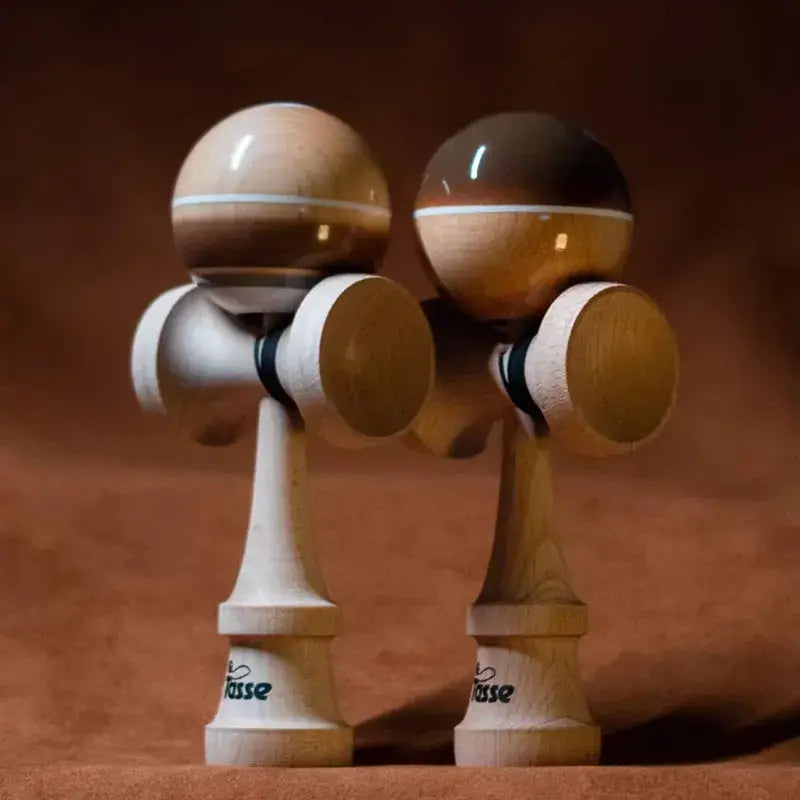 Two Tasse wooden kendamas with glossy natural wood and brown balls, white stripe details, and natural wood handles, displayed side by side on a warm brown background.