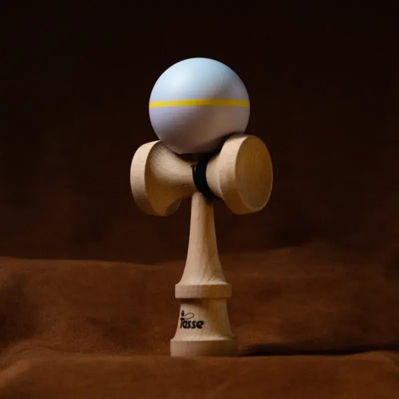 Tasse wooden kendama with a pale lavender ball, yellow stripe detail, and natural wood handle, displayed upright on a warm brown background.