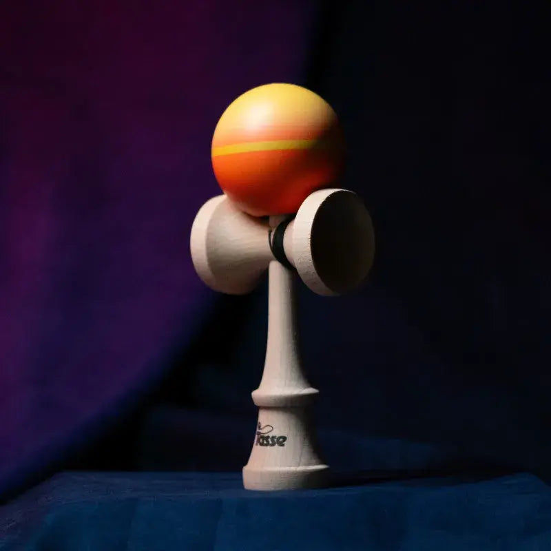 Tasse wooden kendama with an orange-yellow gradient ball and natural wood handle, displayed upright on a dark purple and blue background.