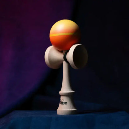 Tasse wooden kendama with an orange-yellow gradient ball and natural wood handle, displayed upright on a dark purple and blue background.
