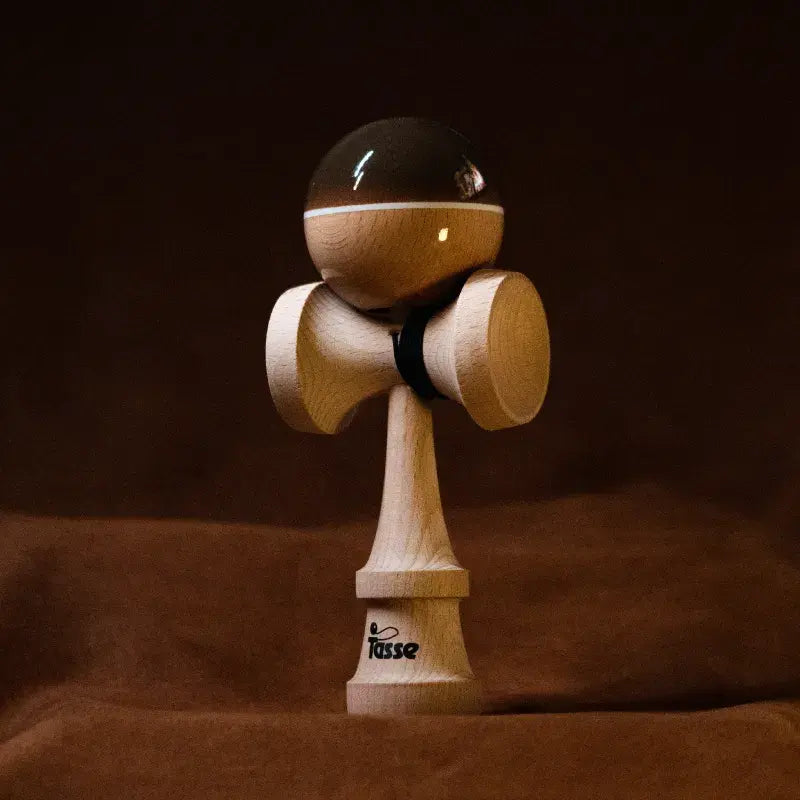 Tasse wooden kendama with a glossy brown and natural wood ball, white stripe detail, and natural wood handle, displayed upright on a warm brown background.