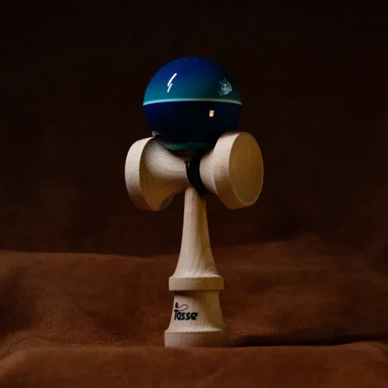 Tasse wooden kendama with a glossy teal and dark blue ball, light stripe detail, and natural wood handle, displayed upright on a warm brown background.