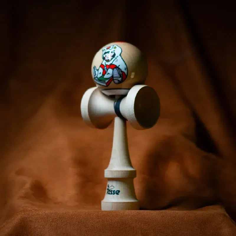 Tasse wooden kendama with a natural wood ball, cartoon character illustration, and natural wood handle, displayed upright on a warm brown background.