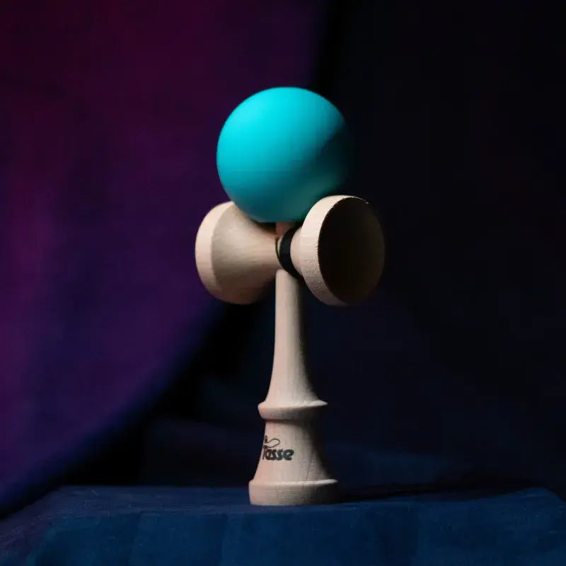 Tasse wooden kendama with a teal ball and natural wood handle, displayed upright on a dark purple and blue background.