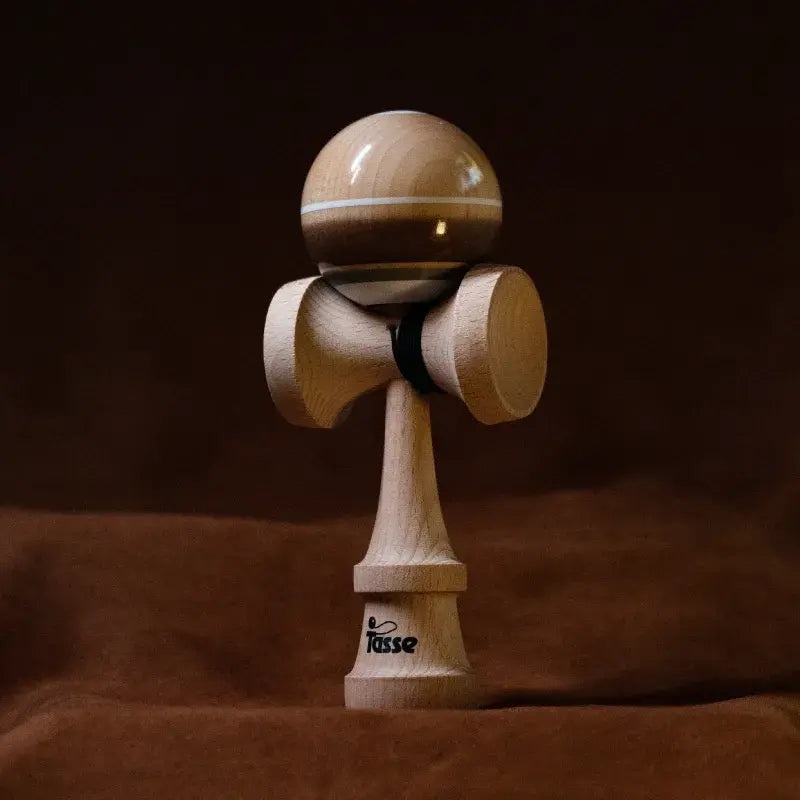 Tasse wooden kendama with a glossy natural wood and brown ball, white stripe detail, and natural wood handle, displayed upright on a warm brown background.