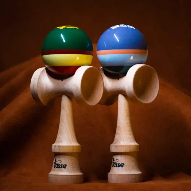 Two Tasse wooden kendamas with glossy multicolor striped balls and natural wood handles, displayed side by side on a warm brown background.