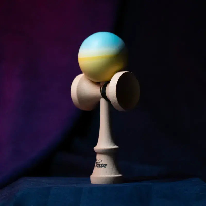 Tasse wooden kendama with a light blue and pale yellow gradient ball and natural wood handle, displayed upright on a dark purple and blue background.