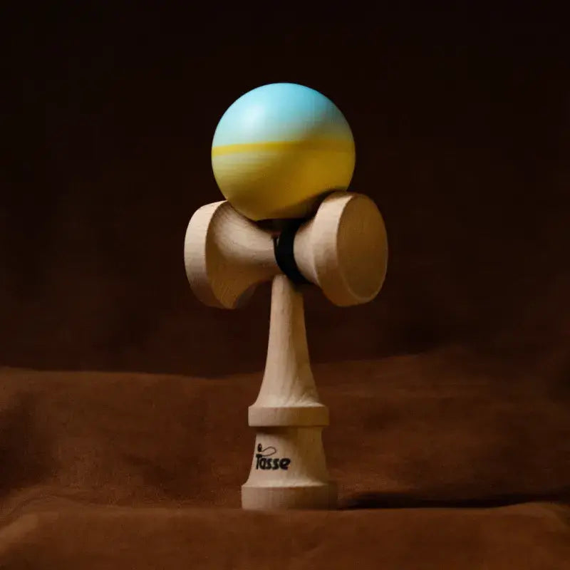 Tasse wooden kendama with a light blue and yellow gradient ball and natural wood handle, displayed upright on a warm brown background.