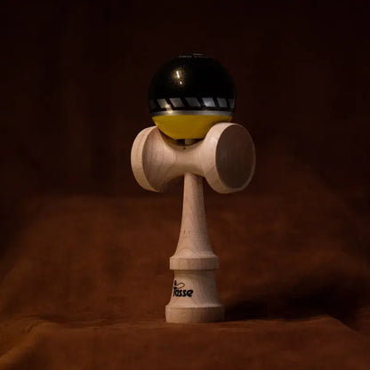 Tasse wooden kendama with a glossy black and yellow ball, gray diagonal stripe detail, and natural wood handle, displayed upright on a warm brown background.