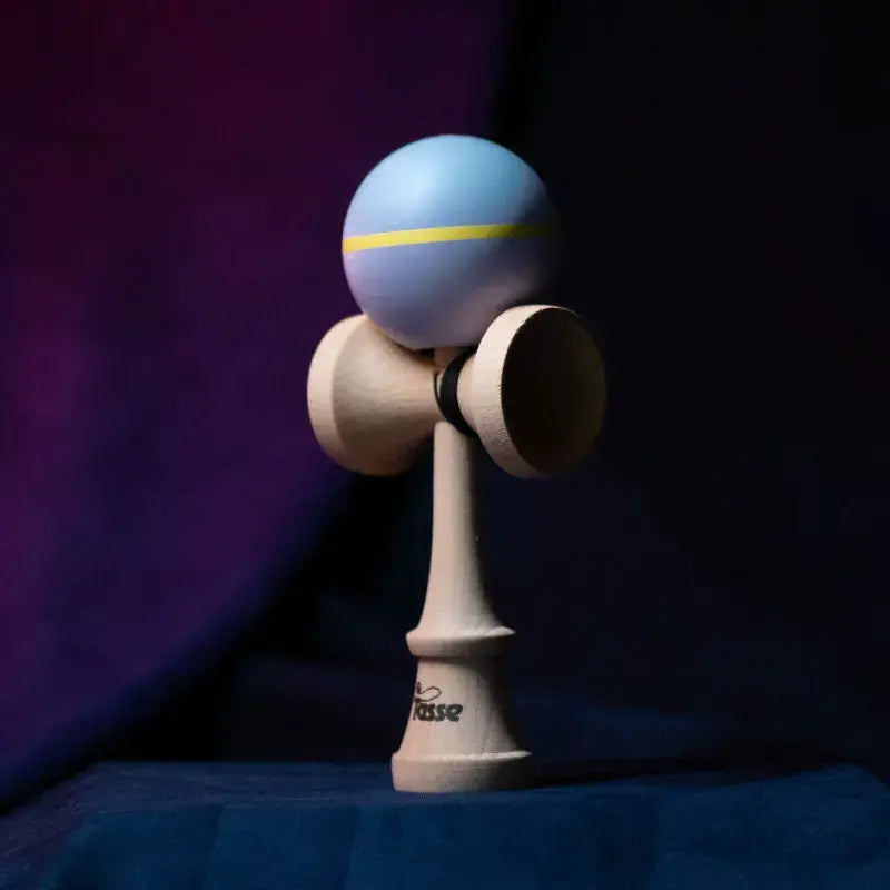 Tasse wooden kendama with a lavender ball, yellow stripe, and natural wood handle, displayed upright on a dark purple and blue background.