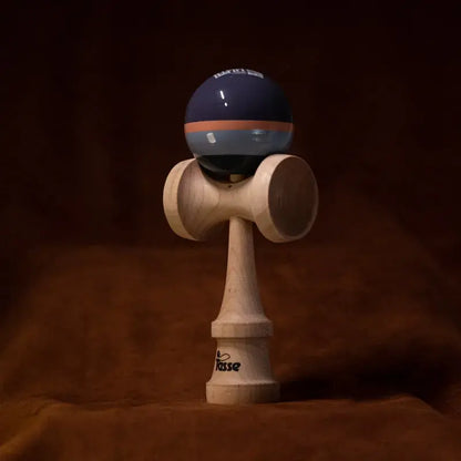 Tasse wooden kendama with a glossy navy ball, orange and blue-gray stripe detail, and natural wood handle, displayed upright on a warm brown background.