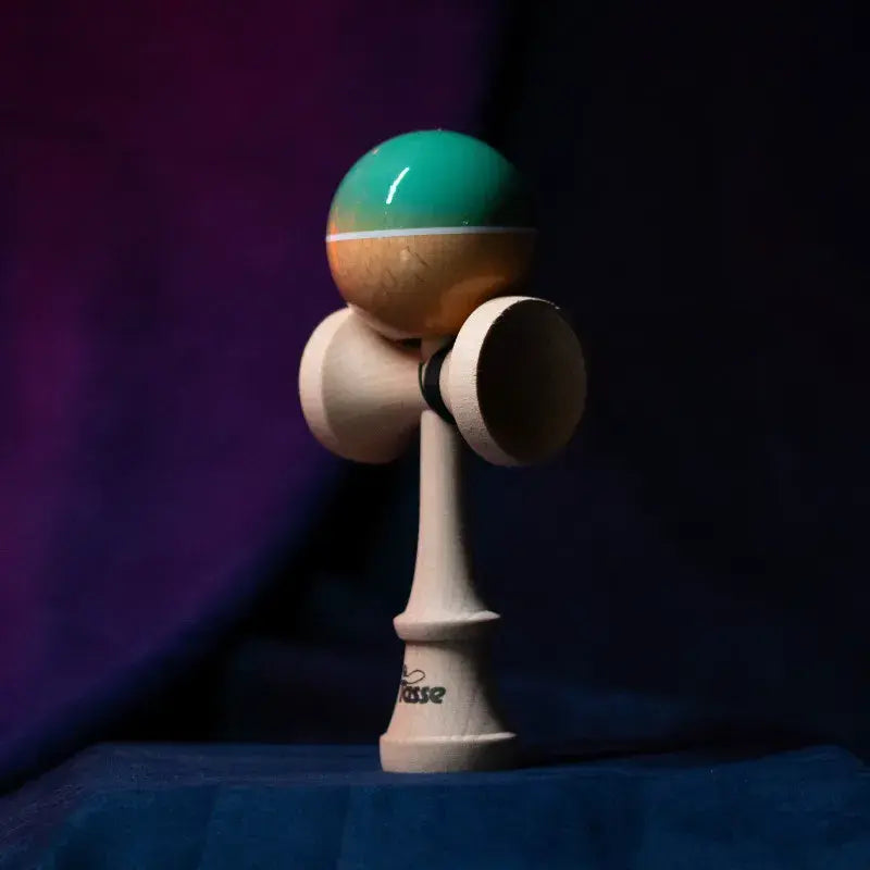 Tasse wooden kendama with a glossy teal and natural wood ball, white stripe detail, and natural wood handle, displayed upright on a dark purple and blue background.