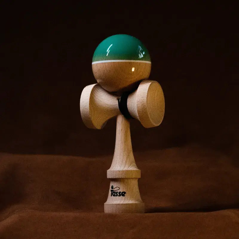 Tasse wooden kendama with a glossy green and natural wood ball, white stripe detail, and natural wood handle, displayed upright on a warm brown background.