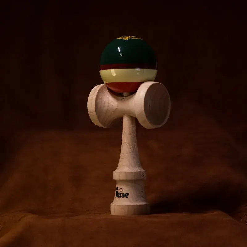 Tasse wooden kendama with a glossy green, cream, and red striped ball and natural wood handle, displayed upright on a warm brown background.