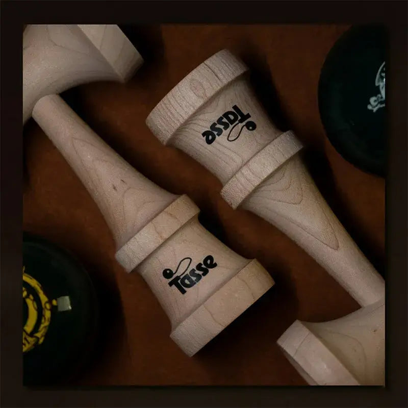 Close-up flat lay of two Tasse wooden kendama handles with logo detail, displayed on a warm brown surface with dark kendama balls partially visible.