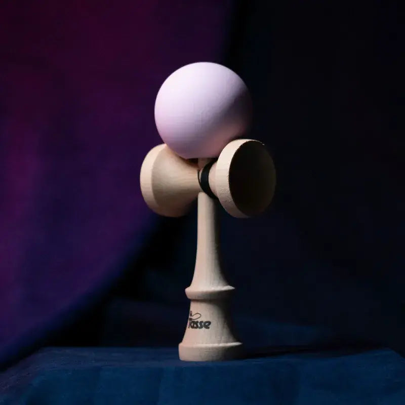 Tasse wooden kendama with a light lavender ball and natural wood handle, displayed upright on a dark purple and blue background.