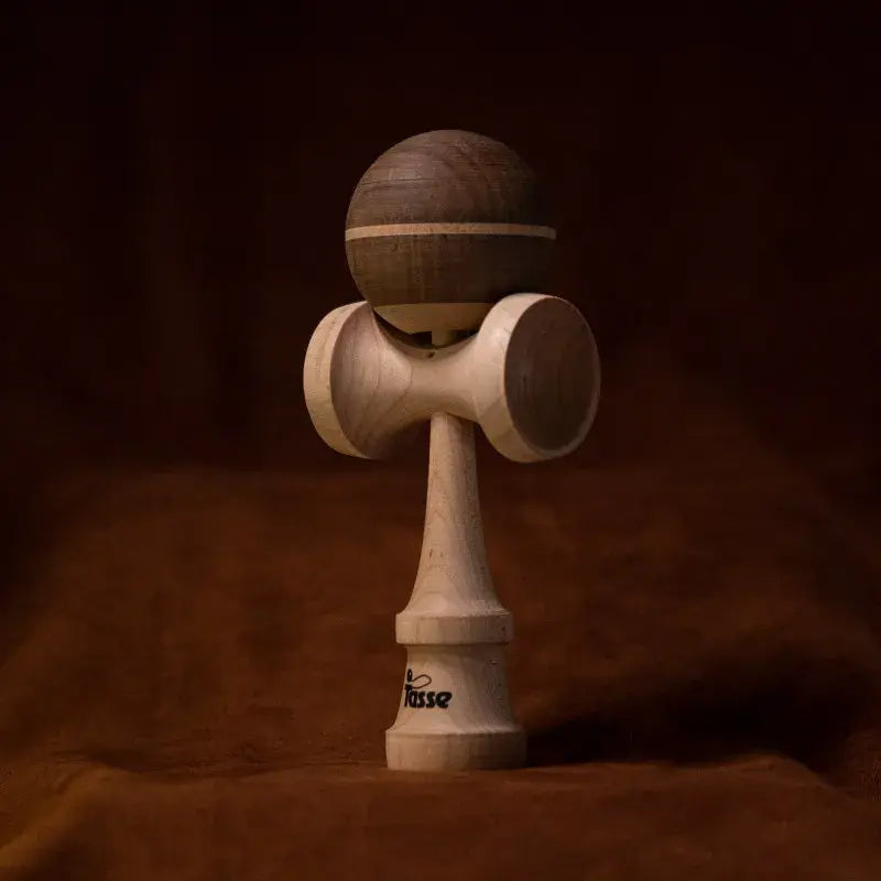 Tasse wooden kendama with a dark wood ball, natural wood stripe detail, and natural wood handle, displayed upright on a warm brown background.