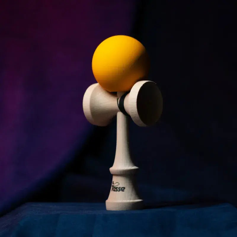 Tasse wooden kendama with a bright yellow ball and natural wood handle, displayed upright on a dark purple and blue background.