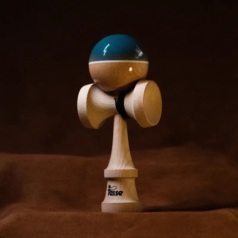Tasse wooden kendama with a glossy blue and natural wood ball, white stripe detail, and natural wood handle, displayed upright on a warm brown background.