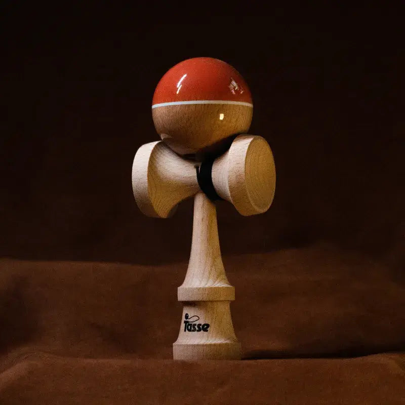 Tasse wooden kendama with a glossy orange and natural wood ball, white stripe detail, and natural wood handle, displayed upright on a warm brown background.
