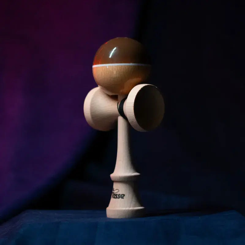 Tasse wooden kendama with a glossy brown and natural wood ball, white stripe detail, and natural wood handle, displayed upright on a dark purple and blue background.