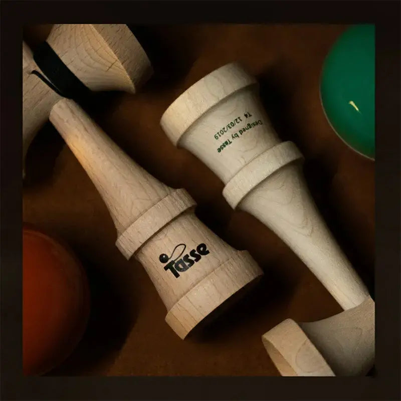 Close-up flat lay of two Tasse wooden kendama handles with logo detail, displayed on a warm brown surface with orange and teal balls partially visible.