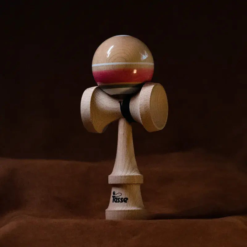 Tasse wooden kendama with a glossy natural wood ball, white stripe, red accent detail, and natural wood handle, displayed upright on a warm brown background.