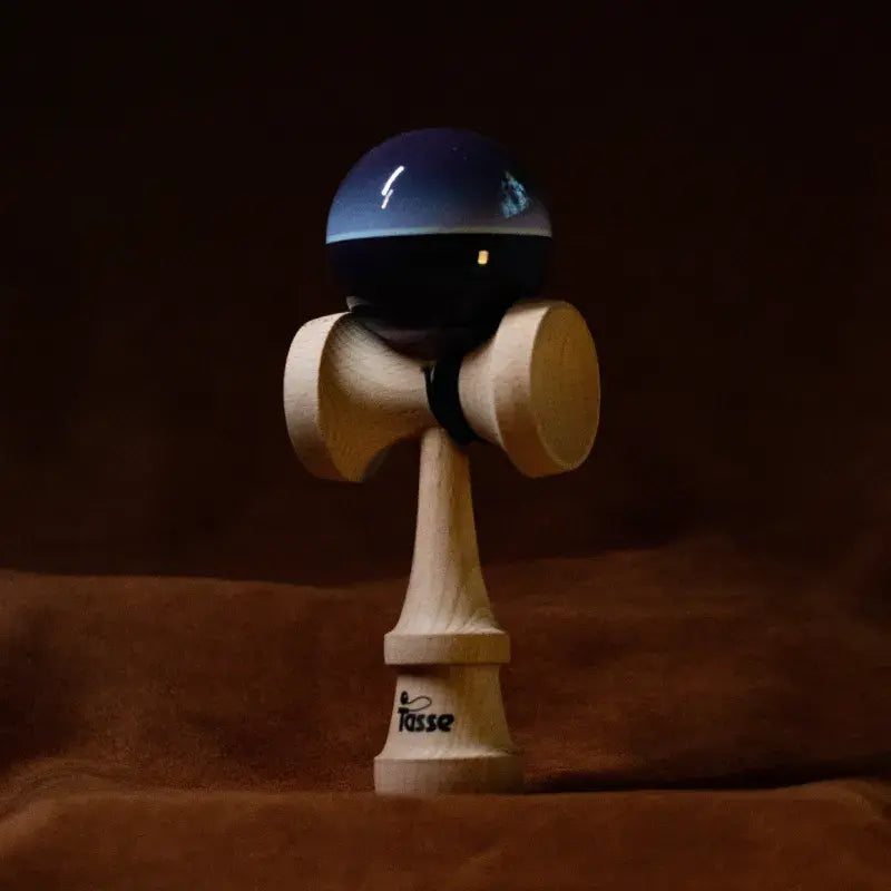 Tasse wooden kendama with a glossy navy and black ball, light blue band detail, and natural wood handle, displayed upright on a warm brown background.