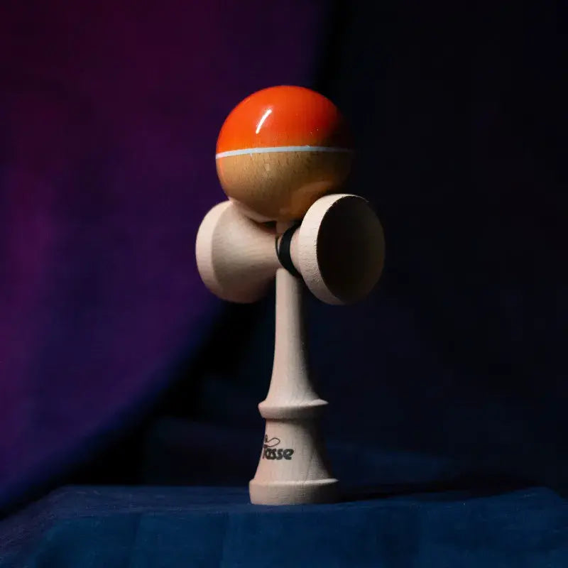 Tasse wooden kendama with a glossy orange and natural wood ball, white stripe detail, and natural wood handle, displayed upright on a dark purple and blue background.