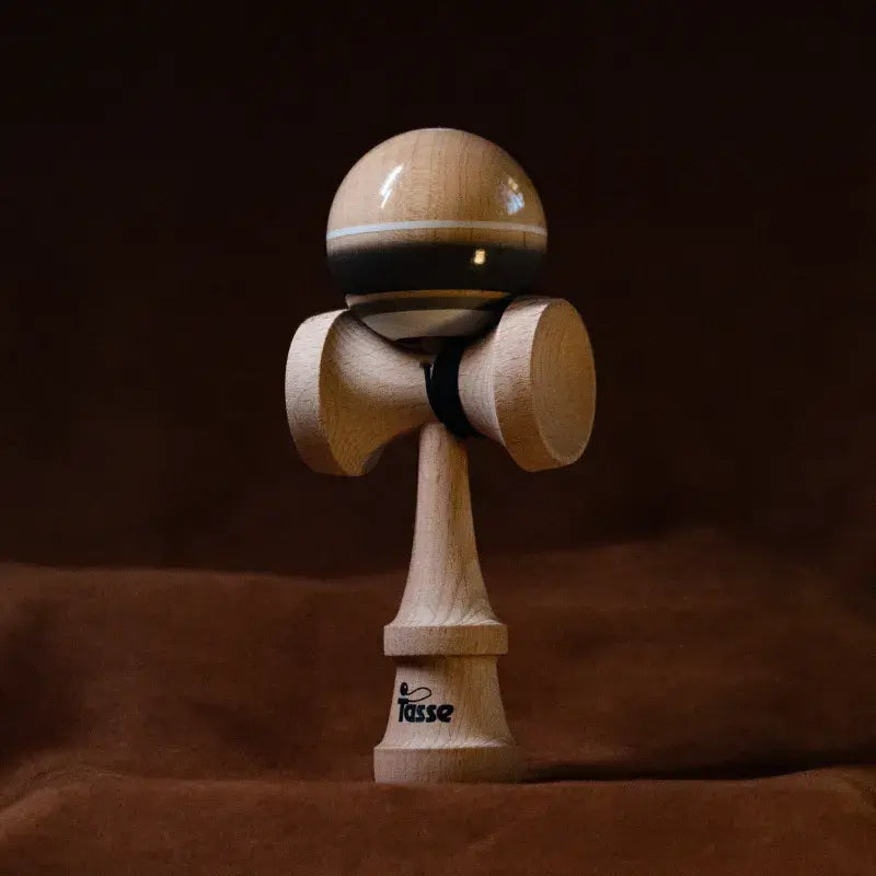 Tasse wooden kendama with a glossy natural wood ball, black band detail, white stripe, and natural wood handle, displayed upright on a warm brown background.