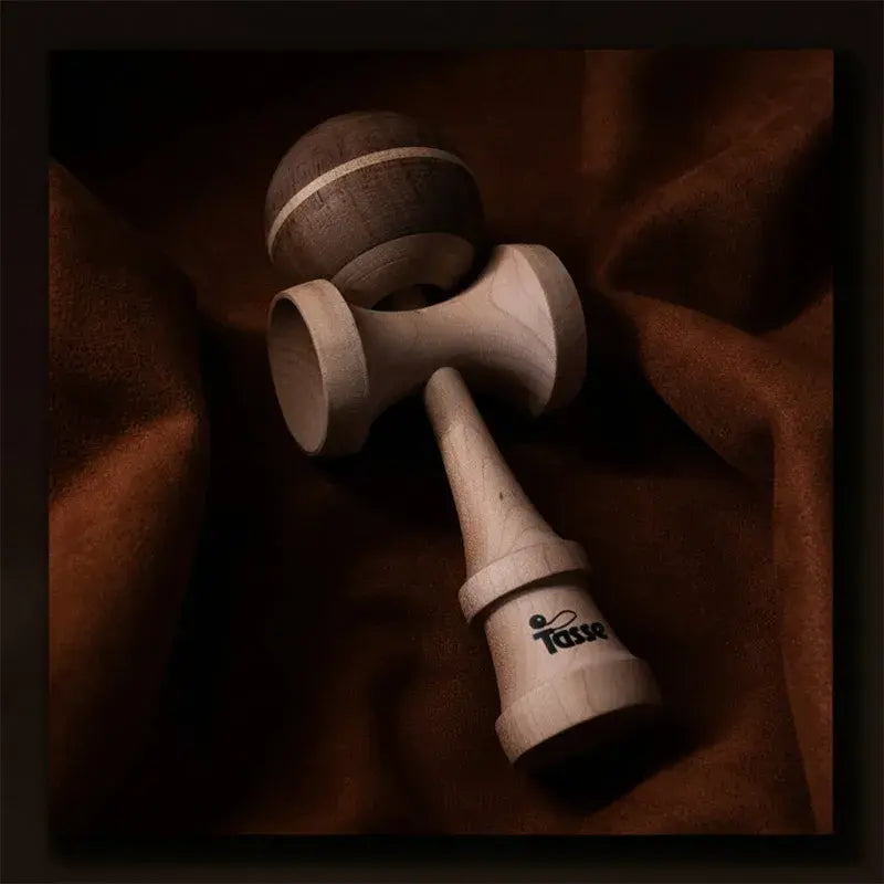 Tasse wooden kendama with a dark wood ball, light stripe detail, and natural wood handle, styled on a warm brown fabric background.