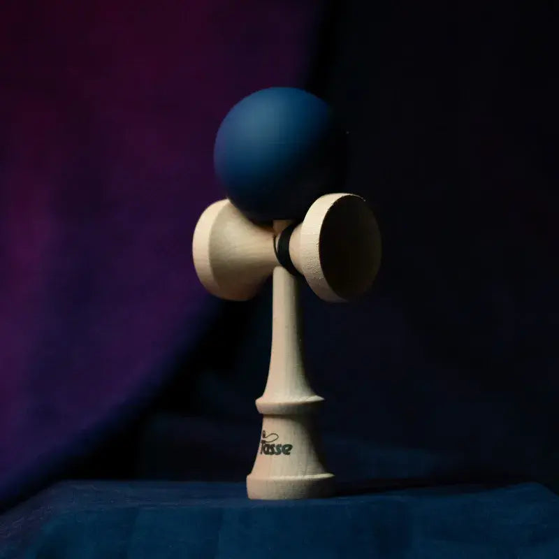 Tasse wooden kendama with a dark blue ball and natural wood handle, displayed upright on a dark purple and blue background.