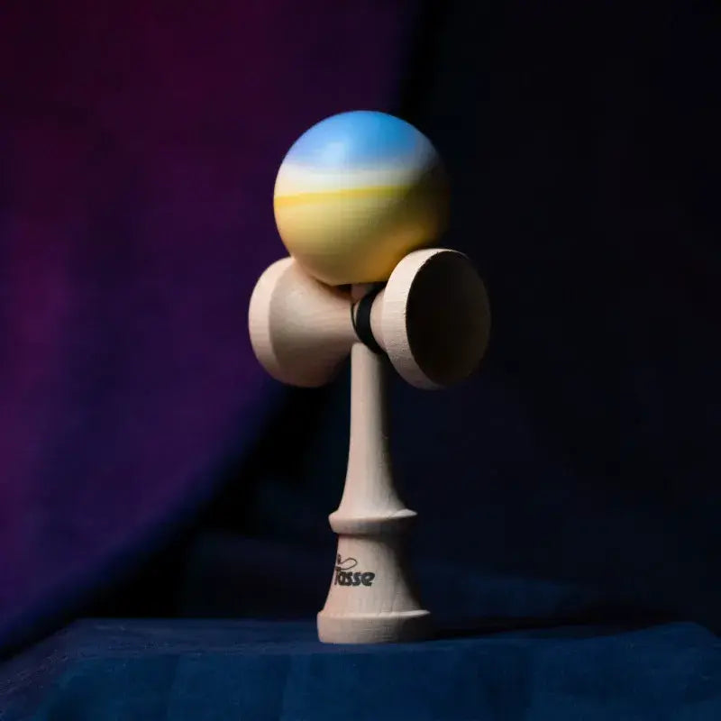 Tasse wooden kendama with a light blue and pale yellow gradient ball and natural wood handle, displayed upright on a dark purple and blue background.
