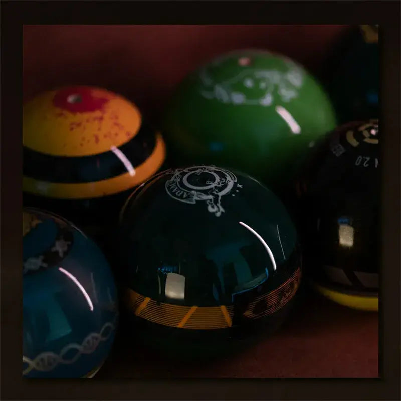 Close-up of glossy Tasse kendama balls in green, blue, yellow, and black finishes with printed graphic details, displayed together on a dark warm-toned surface.