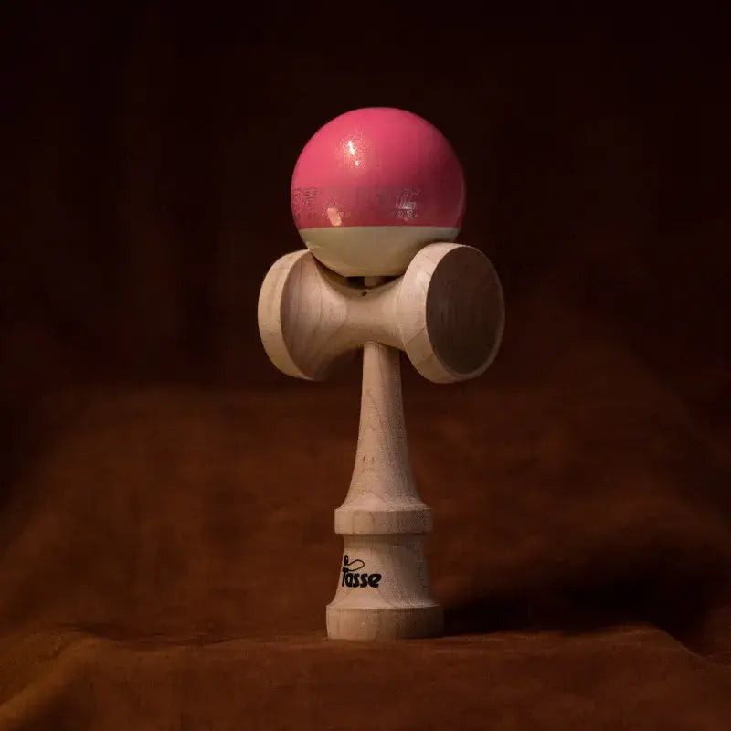 Tasse wooden kendama with a glossy pink and white ball and natural wood handle, displayed upright on a warm brown background.