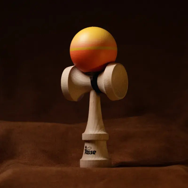 Tasse wooden kendama with an orange and yellow gradient ball and natural wood handle, displayed upright on a warm brown background.