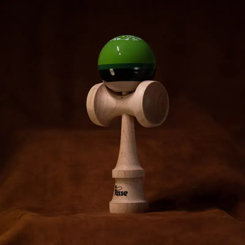 Tasse wooden kendama with a glossy green and black ball and natural wood handle, displayed upright on a warm brown background.