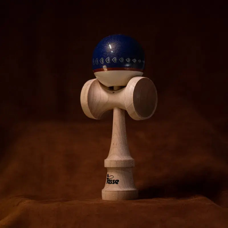 Tasse wooden kendama with a glossy blue ball, white lower section, geometric band detail, and natural wood handle, displayed upright on a warm brown background.