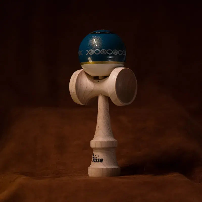Tasse wooden kendama with a glossy blue ball, white lower section, yellow stripe, chain-style graphic detail, and natural wood handle, displayed upright on a warm brown background.