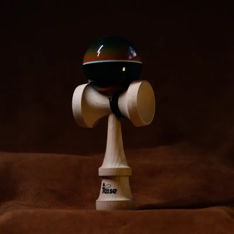 Tasse wooden kendama with a glossy black ball, orange gradient detail, white stripe, and natural wood handle, displayed upright on a warm brown background.