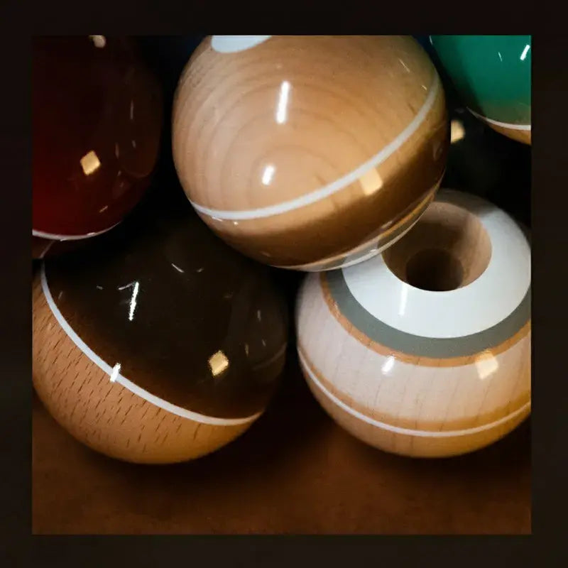 Close-up of glossy Tasse kendama balls in natural wood, brown, and green finishes, with white stripe details and one hole visible, displayed on a warm brown surface.