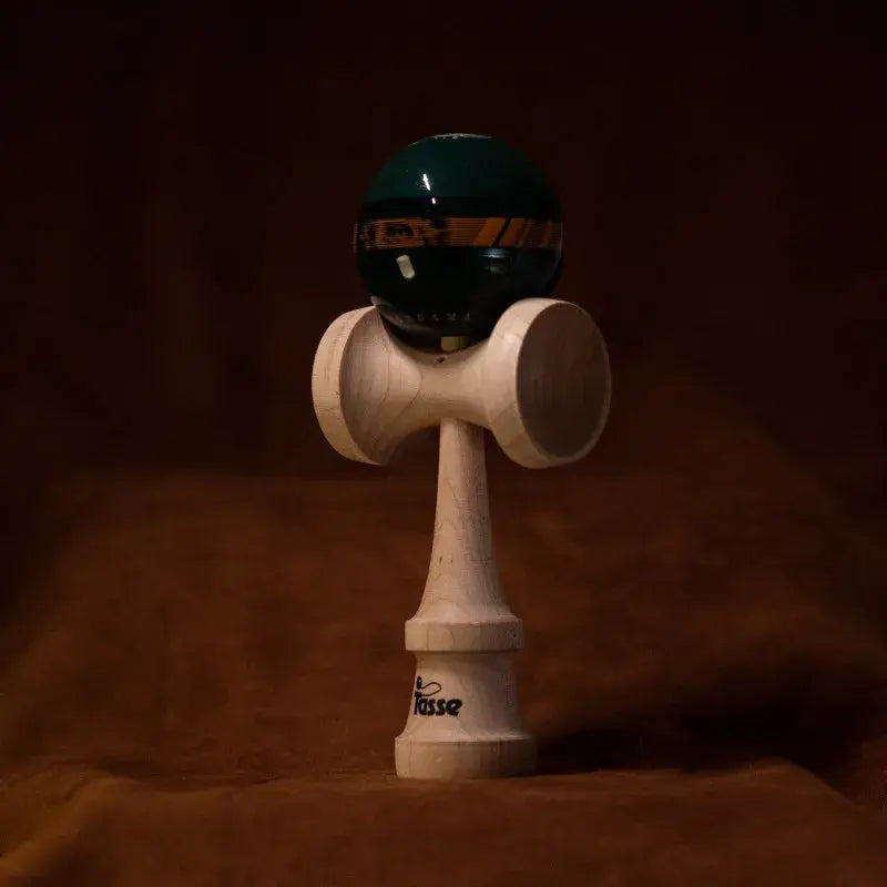 Tasse wooden kendama with a glossy dark green and black ball, gold graphic band detail, and natural wood handle, displayed upright on a warm brown background.