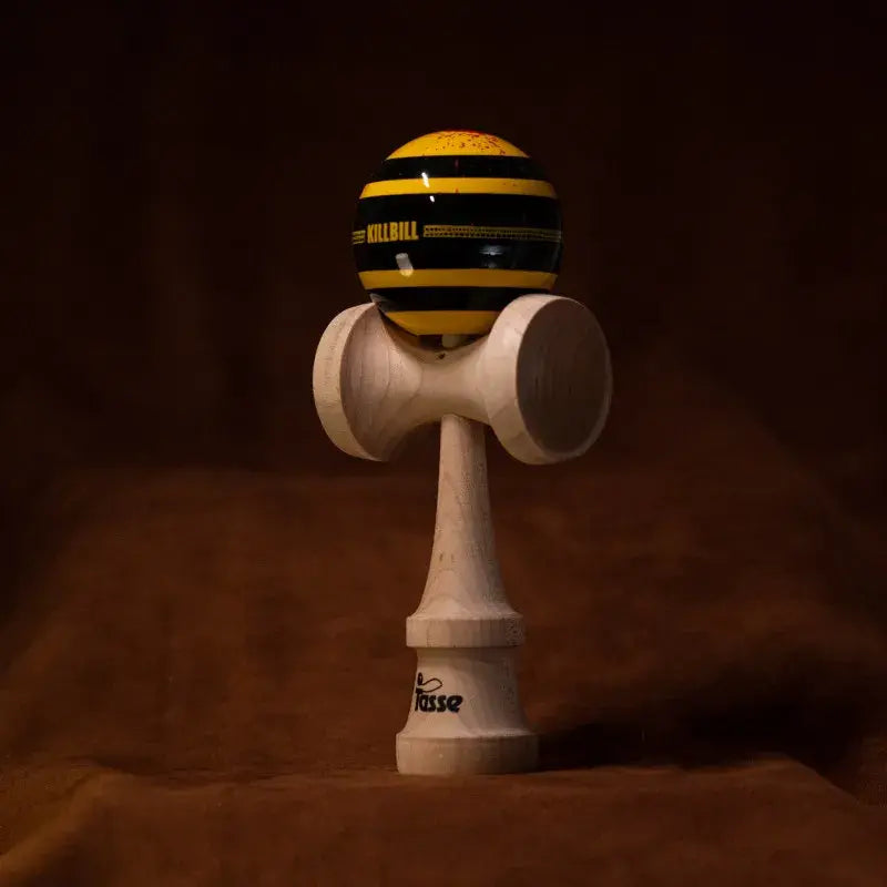 Tasse wooden kendama with a glossy yellow and black striped ball, “Kill Bill” graphic detail, and natural wood handle, displayed upright on a warm brown background.
