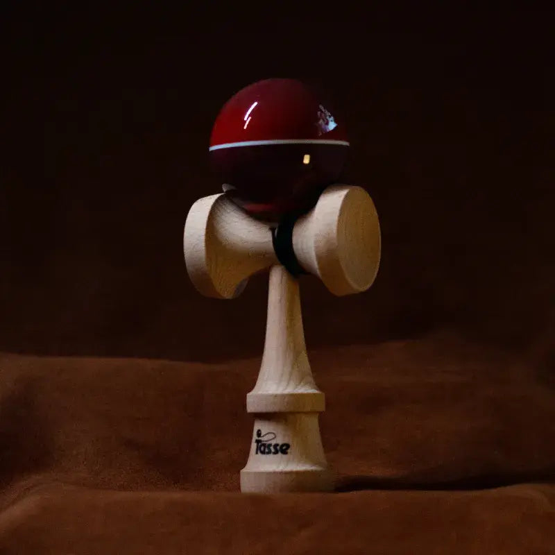 Tasse wooden kendama with a glossy deep red ball, white stripe detail, and natural wood handle, displayed upright on a warm brown background.