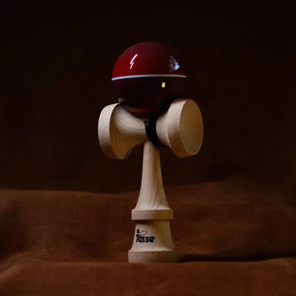 Tasse wooden kendama with a glossy deep red ball, white stripe detail, and natural wood handle, displayed upright on a warm brown background.