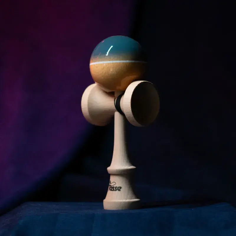 Tasse wooden kendama with a glossy blue-gray and natural wood ball, white stripe detail, and natural wood handle, displayed upright on a dark purple and blue background.