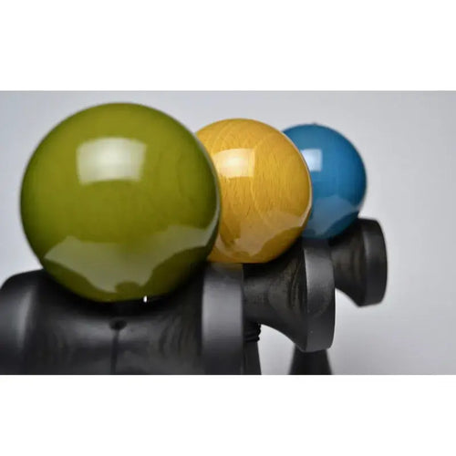 Close-up of three kendamas with glossy olive green, mustard yellow, and blue balls on black wooden handles, displayed against a light background.
